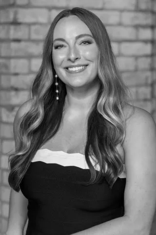 Black and white professional portrait of Jessi Pallan, Creative Director of Iron Diamond Media, wearing a strapless dress with long, wavy hair and pearl earrings, smiling in front of a brick wall.