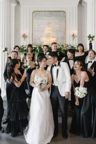 Wedding party featuring an elegant, classic bride and groom surrounded by bridesmaids in black dresses and groomsmen in tuxedos holding white floral bouquets by Poppy Flowers