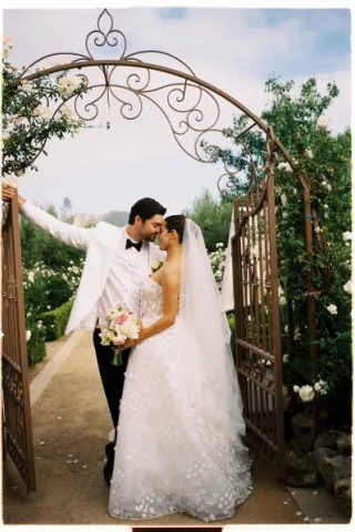 Former Miss California Natasha Alexis Nichelman shares a romantic moment with her groom beneath a vintage wrought-iron gate surrounded by white roses at Tuscan Rose Ranch.