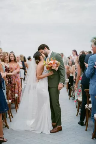 Bride in slip dress, long veil and colorful bouquet, groom in green suit kiss in aisle