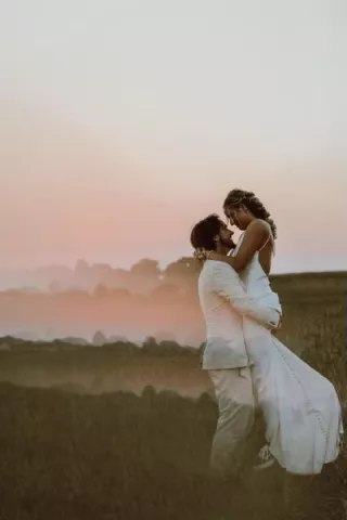 Groom lifts Bride in embrace in a field at sunset