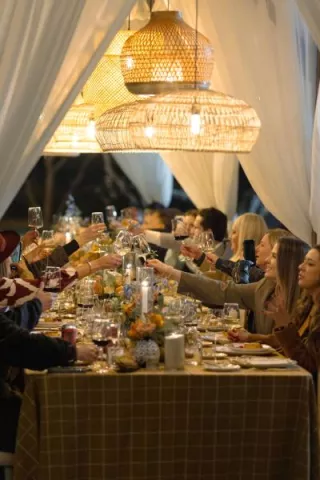 Guests raising wine glasses in a toast under warm, glowing woven chandeliers at a beautifully decorated outdoor dinner table with floral arrangements and candlelight