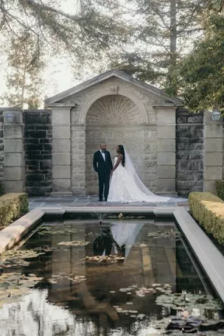 Bride and groom pose by pond, framed by stone architecture at Greystone Mansion