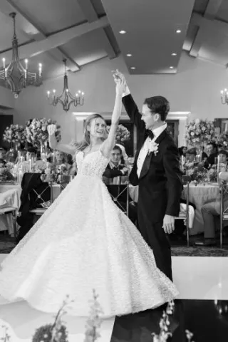 Groom twirls bride on black and white checkered indoor dance floor while their friends and family look on from their tables