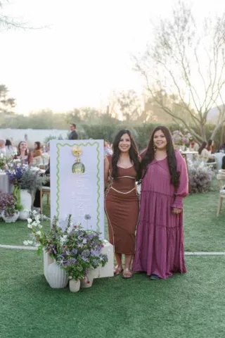 Two stylish women standing next to a modern event welcome sign surrounded by lush purple and green floral arrangements at an outdoor wedding industry gathering.