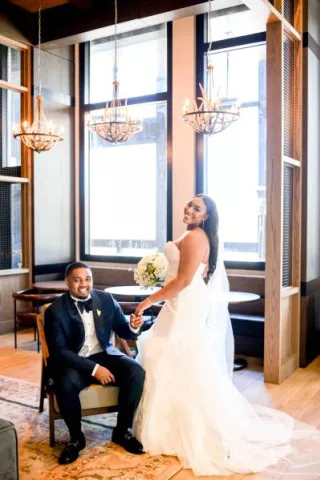 Groom sits and bride stands next to him, posing in the lobby of The Trade Hotel