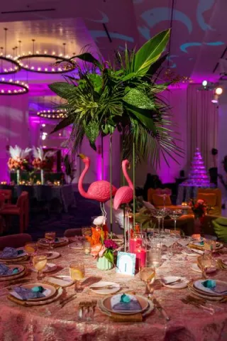 A vibrant dining table at the Curate Gala 2024 features pink flamingo centerpieces, lush tropical greenery, gold flatware, and teal napkins, embodying the Palm Royale theme in the ballroom decor