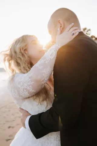 Bride and groom embrace during golden hour on the beach at Rosewood Miramar Beach, bride wearing a lace-sleeved gown and groom in a classic black tuxedo