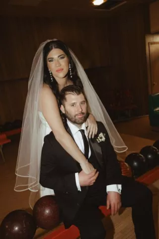 A newlywed couple poses at a vintage bowling alley, exuding confidence and fun. The bride, wearing a classic veil and modern gown, drapes her arms over the groom, who is dressed in a sleek black suit, as bowling balls line the table in front of them.