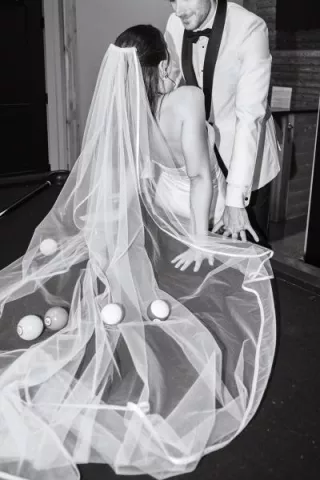 Bride and groom lean against the pool table with pool balls scattered throughout the bride's veil, in black and white