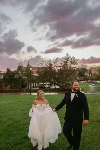Bride and groom walk through field against a stormy sunset