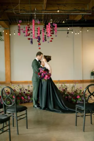 Bride in a black wedding dress and groom in a green suit standing beneath hanging pink and purple flowers at their modern indoor winter wedding ceremony