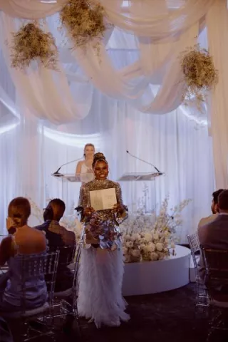 An honoree in a silver sequin top and tiered white tulle skirt holds their award on stage at the Wisconsin Bride Best of 2025 event, framed by cloud-like florals and soft draping decor.