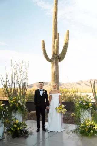 Bride and groom stand hand in hand at the altar, framed by bright blooms and cacti at The Willow Weddings & Event Center