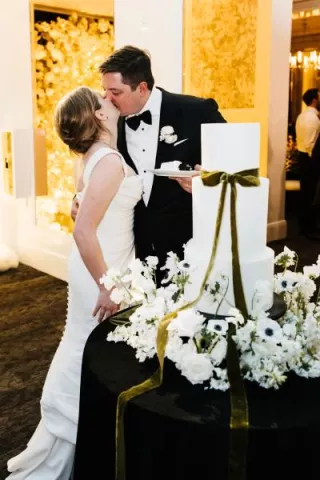 Paige and Johnnie kissing with their cake, surrounded by flowers and a green bow. 