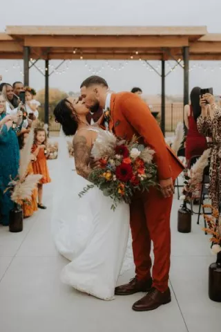 The newlyweds share a kiss down the aisle after their ceremony, with fall-inspired florals, pampas grass and joyful guests in the background.