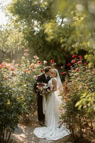Groom kisses bride on the cheek, surrounded by vibrant flowers and bathed in sunlight