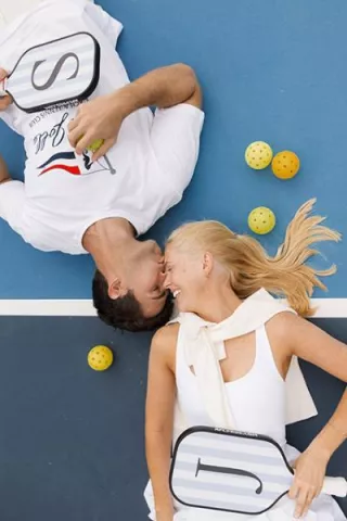 Jennifer and Sean lay on the pickleball court facing each other surrounded by pickleballs