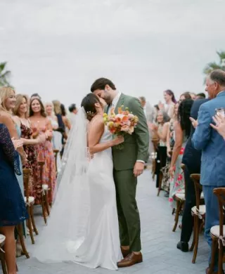 Bride in slip dress, long veil and colorful bouquet, groom in green suit kiss in aisle