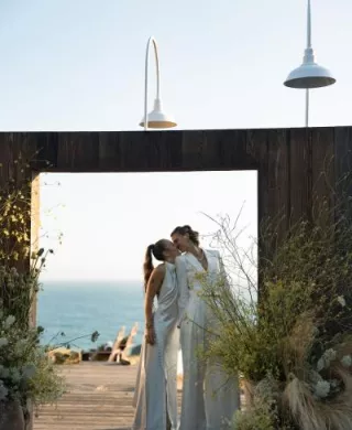 Couple shares a kiss in outdoor door frame, framed by yellow wildflowers and the sea in the background at The Sea Ranch Lodge
