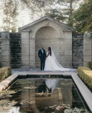 Bride and groom pose by pond, framed by stone architecture at Greystone Mansion