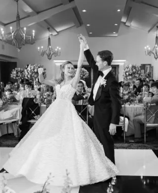 Groom twirls bride on black and white checkered indoor dance floor while their friends and family look on from their tables