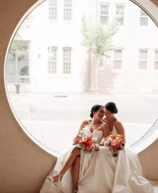 Caroline and Amy cozy up against a large circular window on their wedding day at Overture Center for the Arts