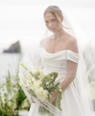 Model poses with a bouquet of lush greenery in an off the shoulder gown and veil with the coast of Crook Point in the background