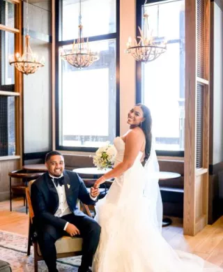 Groom sits and bride stands next to him, posing in the lobby of The Trade Hotel