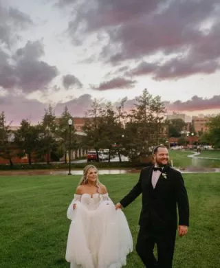 Bride and groom walk through field against a stormy sunset