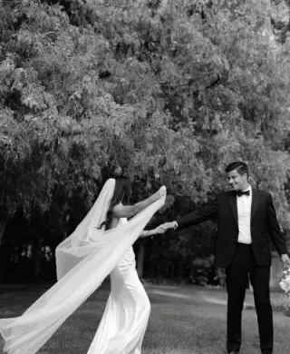 Black and white portrait of bride approaching her groom, her long veil trailing behind her