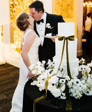 Paige and Johnnie kissing with their cake, surrounded by flowers and a green bow. 
