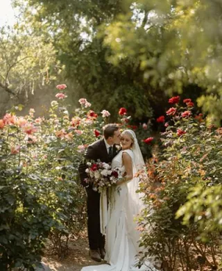 Groom kisses bride on the cheek, surrounded by vibrant flowers and bathed in sunlight