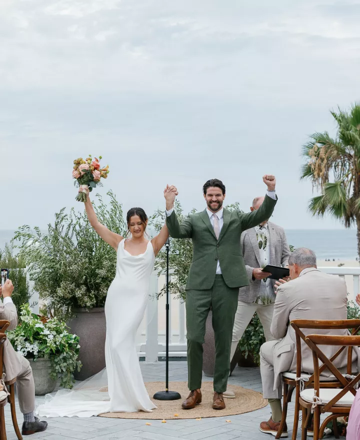 Bride in sleek slip gown and groom in green suit at altar on patio overlooking Santa Monica beach