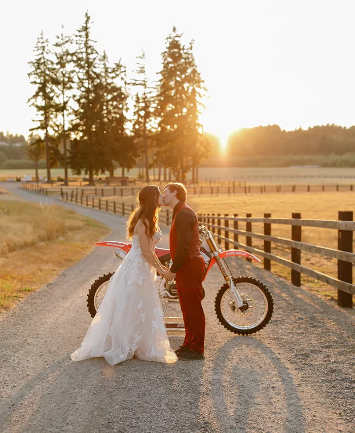 Bride in elegant gown and groom in red suit kiss at sunset with dirt bike in background