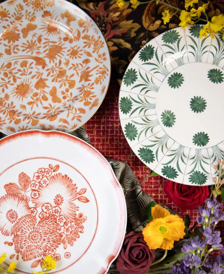 Three designer plates from Maison de Carine—orange Sacred Bird and Butterfly, green Topiary, and coral Oscar’s Coral—styled with roses, poppies, and patterned linens