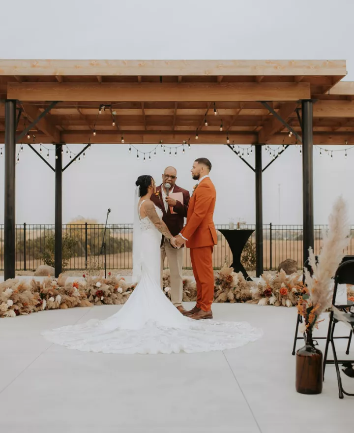 Bride and groom hold hands at the altar during their outdoor fall wedding ceremony, surrounded by pampas grass and warm-toned florals under a wooden pergola.