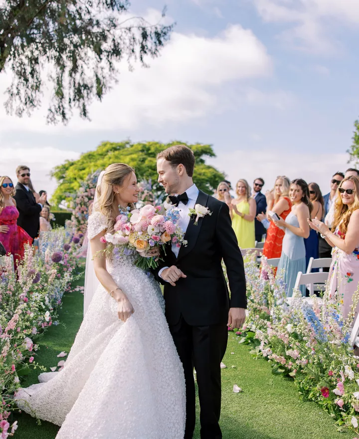 Bride and groom walk back down outdoor floral lined aisle after getting married at La Jolla Country Club