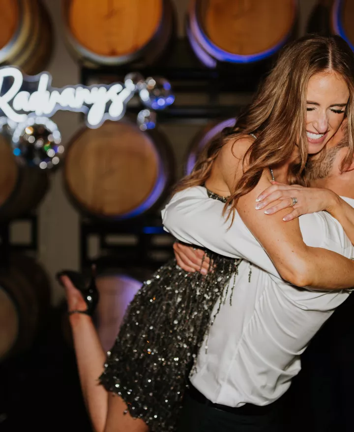 Bride and groom happily embrace in their festive after party outfits in front of a neon sign and wine barrels at JM Cellars