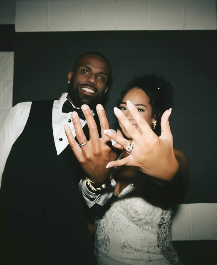 Bride and groom flaunt their rings, holding their hands up as they stand against a black and white checkered background