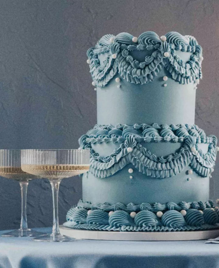 Dusty blue styled scene featuring two-tiered lambeth cake, candles and two glasses of champagne on silky tablecloths