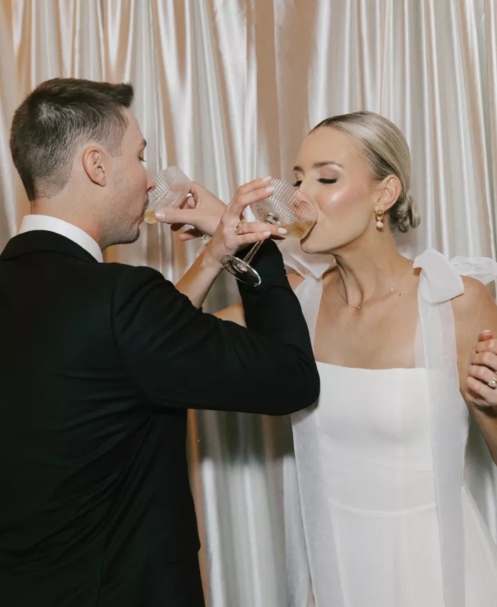 Bride and groom intertwine arms, sipping champagne against a draped backdrop 