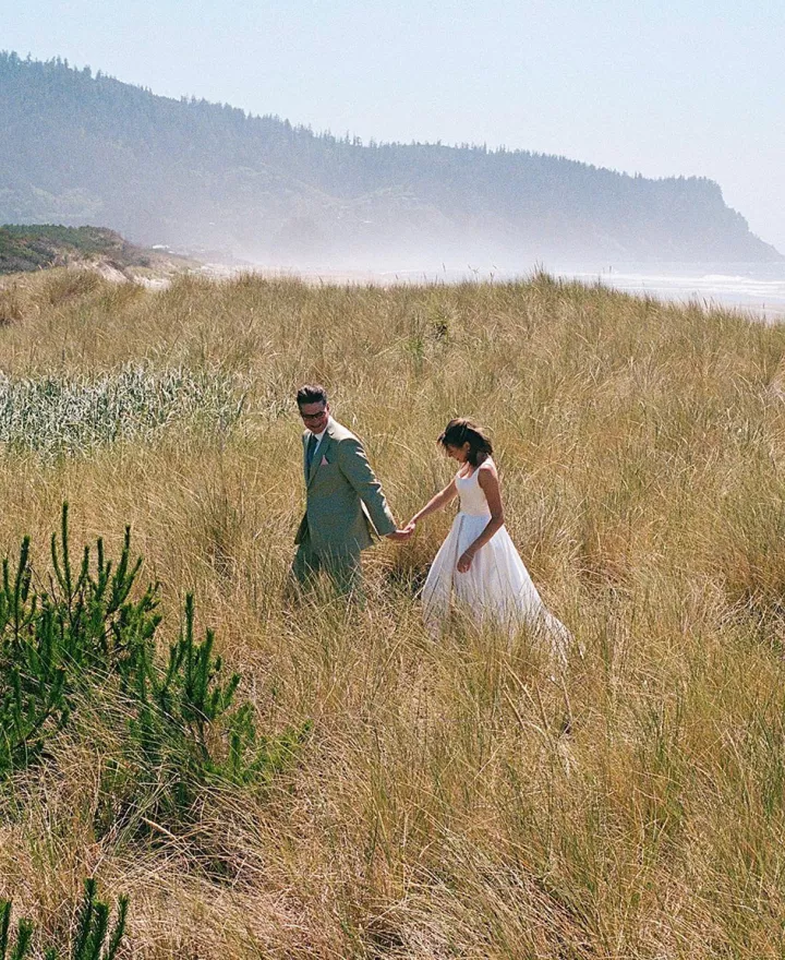Bride and groom walk hand in hand through oceanside field in coastal wedding day