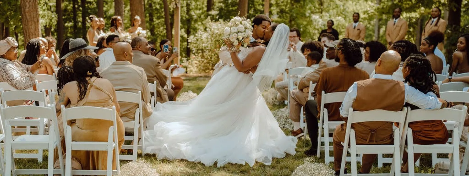 Outdoor wedding ceremony featuring a bride and groom sharing a kiss, surrounded by guests in a lush forest setting, with floral arrangements by Poppy Flowers