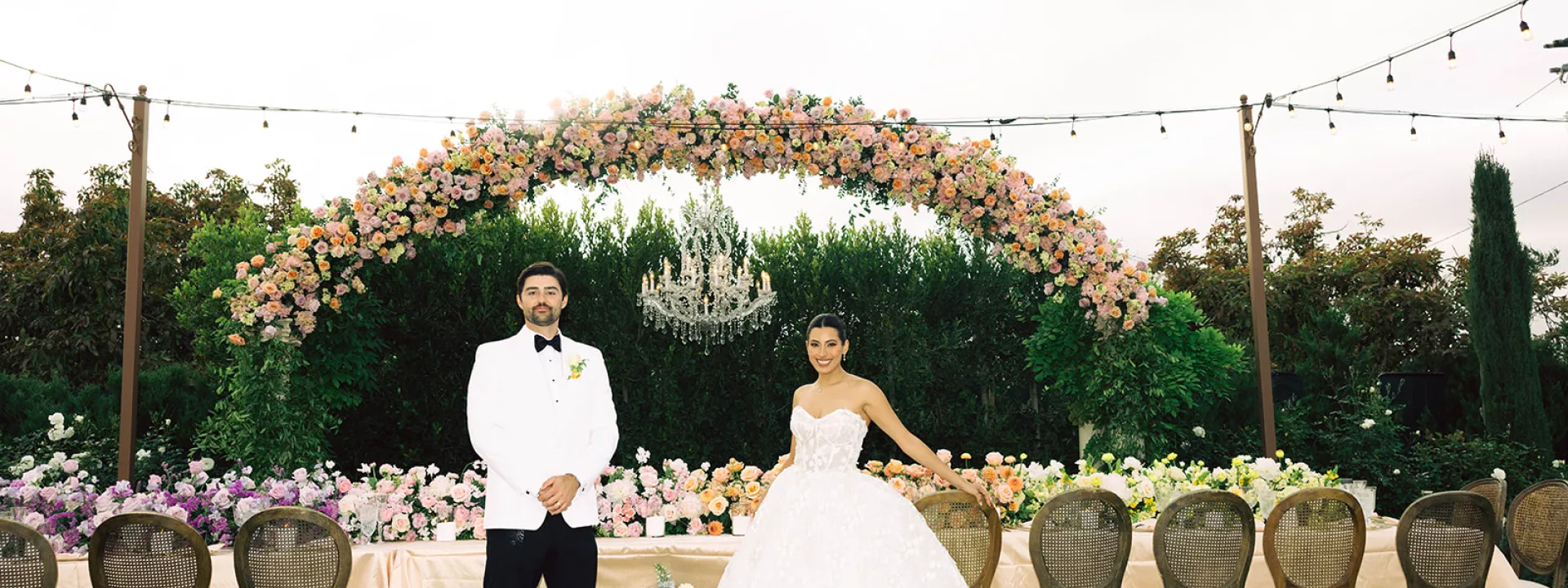 Former Miss California Natasha Alexis Nichelman and Zach Nichelman pose at their reception's head table under a 15-foot floral arch with cascading flowers and a chandelier at Tuscan Rose Ranch.
