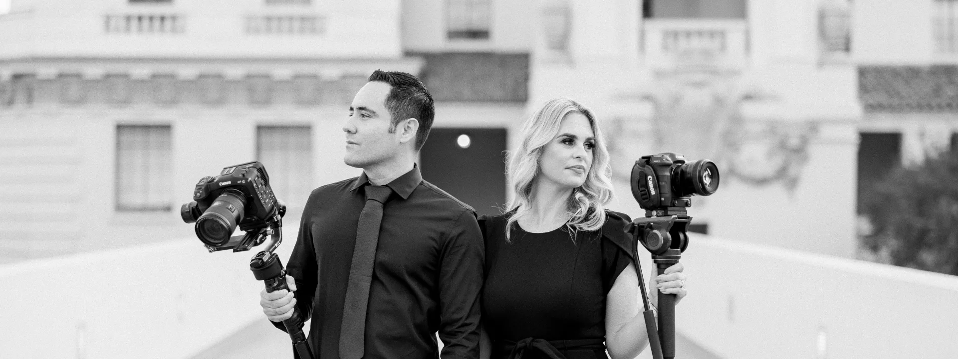 Professional videography team Richard Saenz and Tracy Saenz of Royal Films holding cameras and standing confidently in black attire on a bridge in front of a historic building