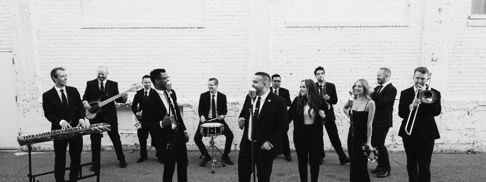 Black-and-white photo of the Downtown Collective band performing outdoors with a full ensemble, including vocalists, keyboard, brass, and percussion
