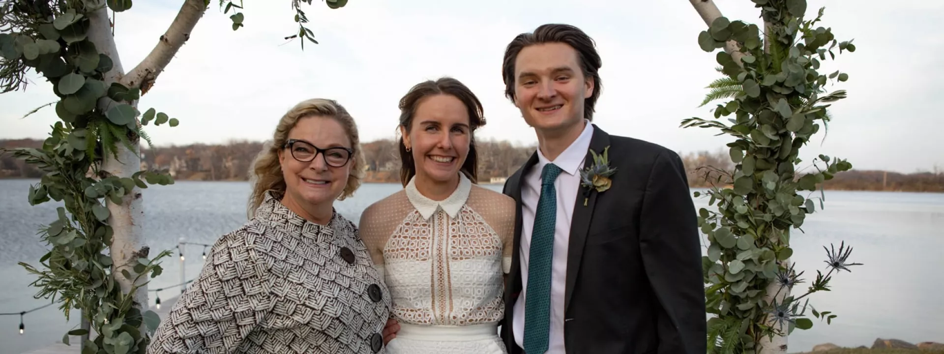 Wedding officiant Dawn Dacut of Flutterby Celebrations with a smiling bride and groom under a birchwood arbor adorned with greenery by a lake