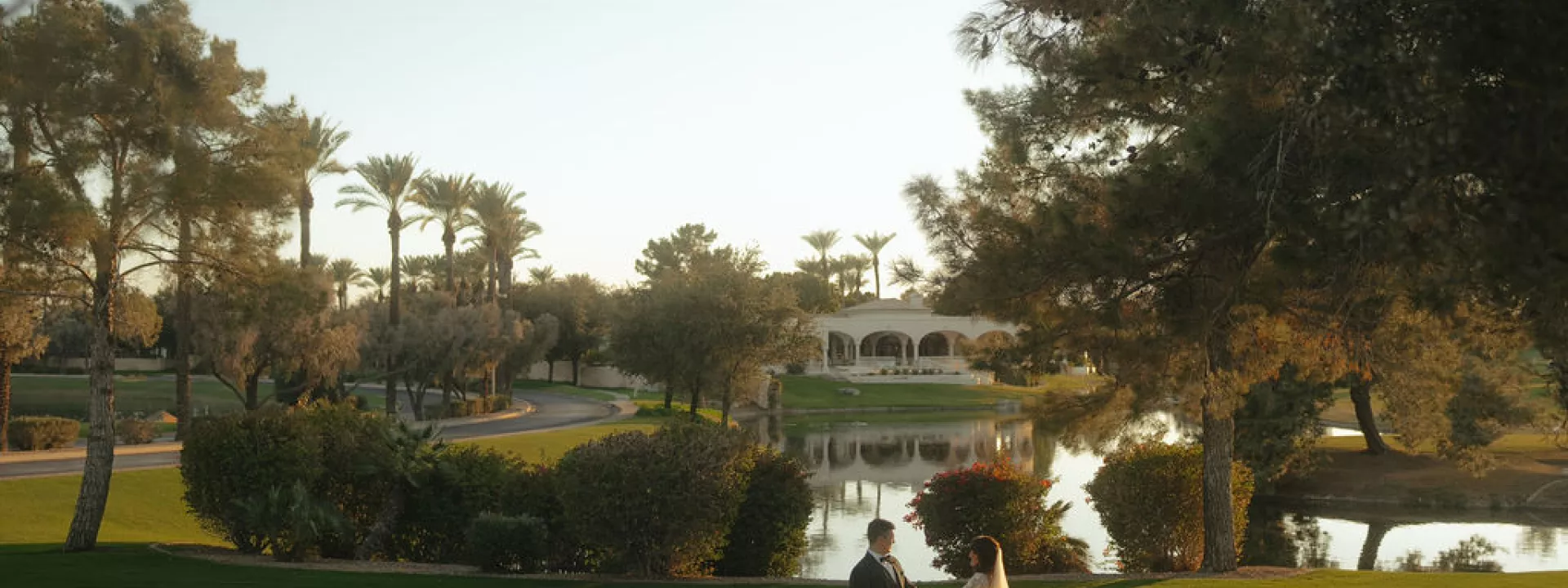A bride and groom share an intimate moment on the lush green lawn of Ocotillo Golf Club in Arizona, with a picturesque lake, palm trees, and a Spanish-style clubhouse in the background at sunset. 