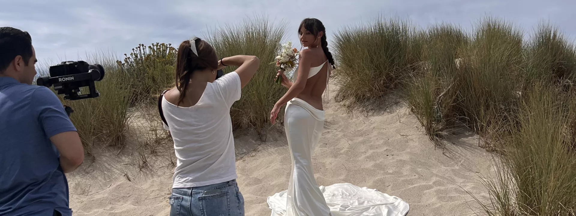 Behind-the-scenes photo of a bridal fashion shoot on a sandy beach, with a model in a satin gown posing in front of tall dune grass, captured by a photographer and videographer in natural light.